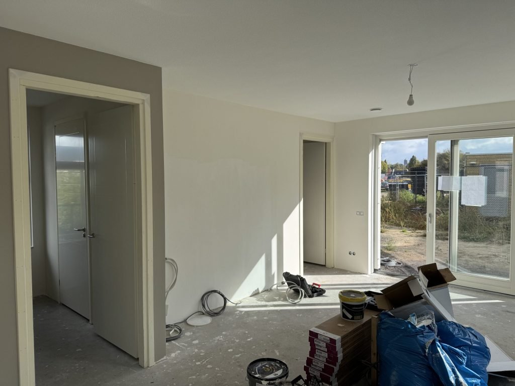 Woonkamer van een net opgeleverd huis met kale wanden, open binnendeuren en het bouwterrein achter de pui